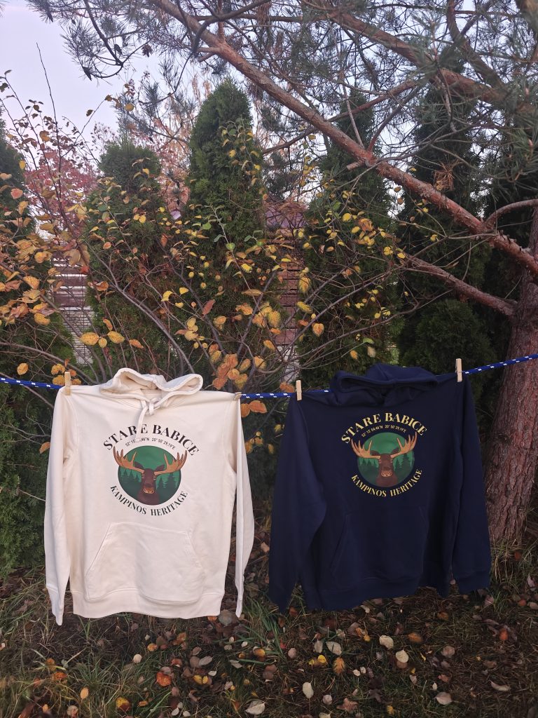 Dwie bluzy „Stare Babice – Kampinos Heritage” wiszące na sznurku w ogrodzie: beżowa i granatowa, z widocznym dużym nadrukiem łosia.